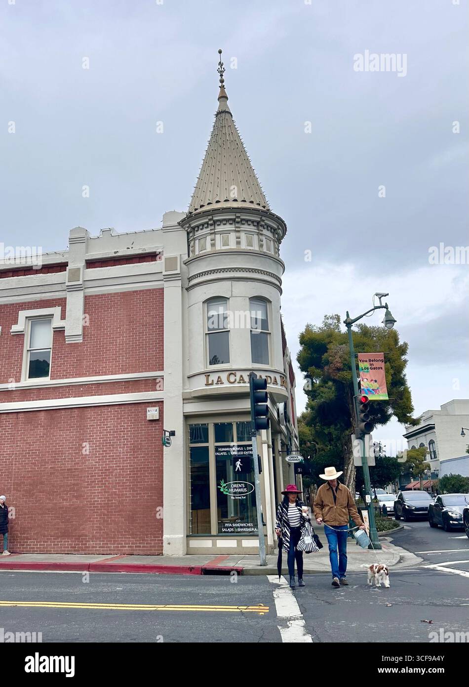 Persone che camminano con un cane dall'altra parte della strada vicino a uno storico edificio in mattoni nel centro di Los Gatos con vetrine e striscioni stradali - Immagine stock catturata con smartphone
