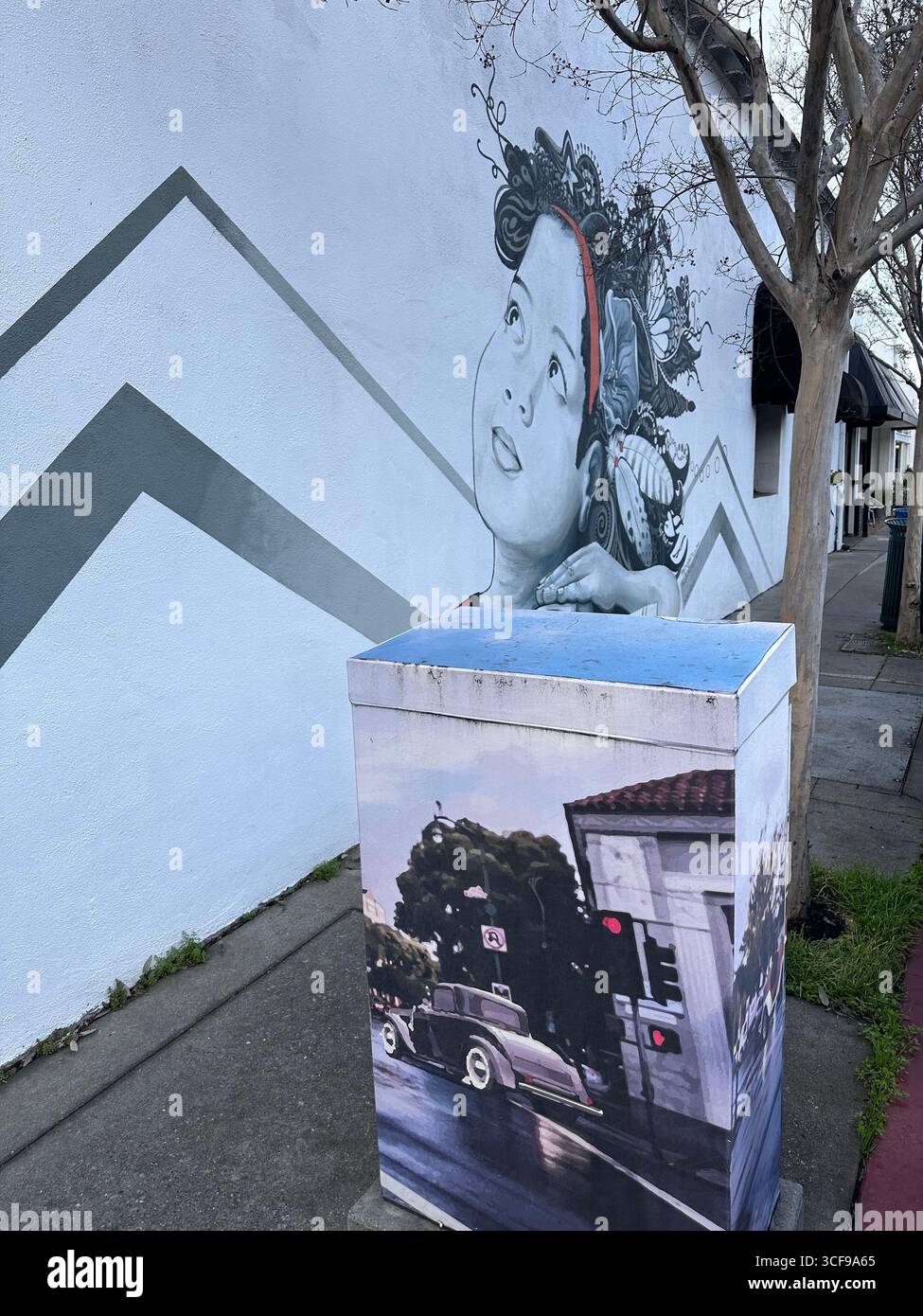 Un murale di strada che raffigura un bambino con elaborati disegni di capelli accanto a una cassetta dipinta che mostra un'auto d'epoca e la scena di un edificio storico Foto Stock