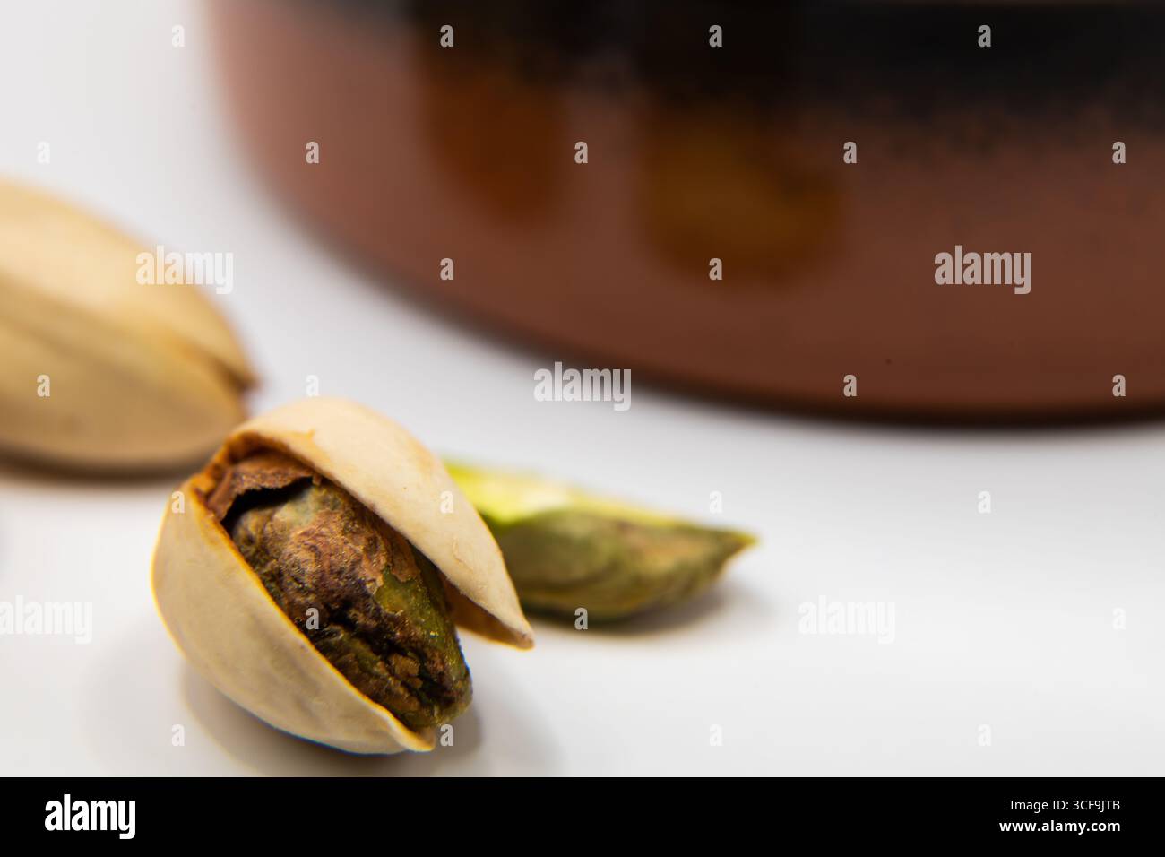 Macro primo piano di un singolo dado di pistacchio su una superficie bianca, con un piatto di terracotta sfocato di noci sullo sfondo, evidenziando e mangiando sano Foto Stock
