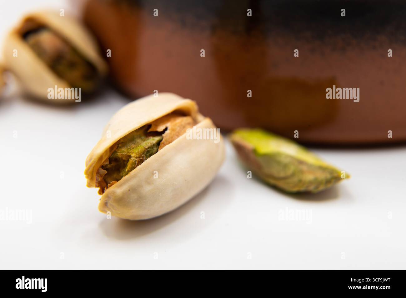 Macro primo piano di un singolo dado di pistacchio su una superficie bianca, con un piatto di terracotta sfocato di noci sullo sfondo, evidenziando e mangiando sano Foto Stock