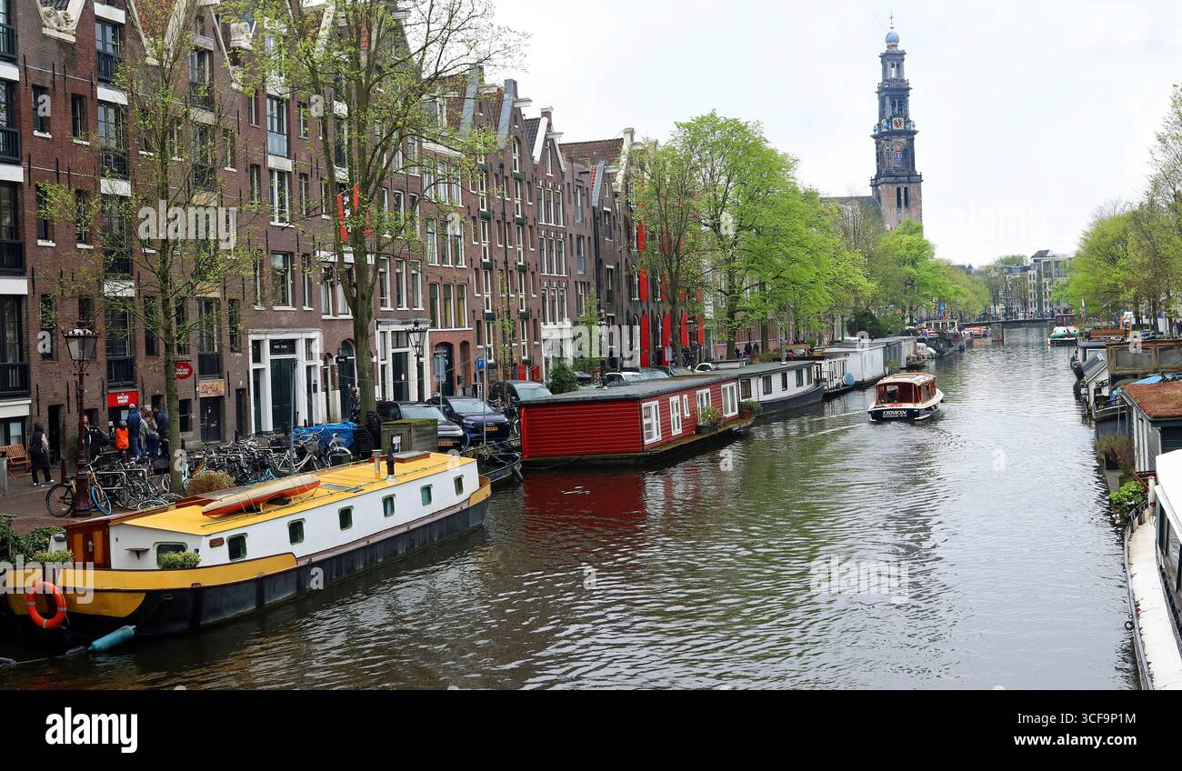 Una piccola barca scende lungo un canale fiancheggiato da chiatte per case galleggianti. La chiesa di Westerkerk può essere vista in lontananza. Aprile ad Amsterdam, Paesi Bassi, Europa Foto Stock Una piccola barca scende lungo un canale fiancheggiato da chiatte per case galleggianti. La chiesa di Westerkerk può essere vista in lontananza. Aprile ad Amsterdam, Paesi Bassi, Europa Foto Stock