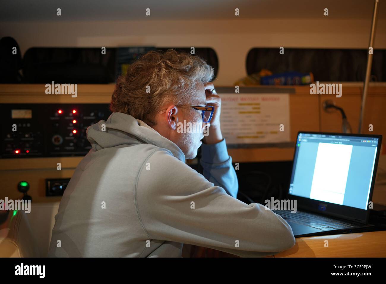 Uomo concentrato che indossa occhiali che lavora su un computer portatile nella cabina di yacht di notte, concetto di lavoro a distanza, stile di vita a vela e nomade digitale a Tenerif Foto Stock