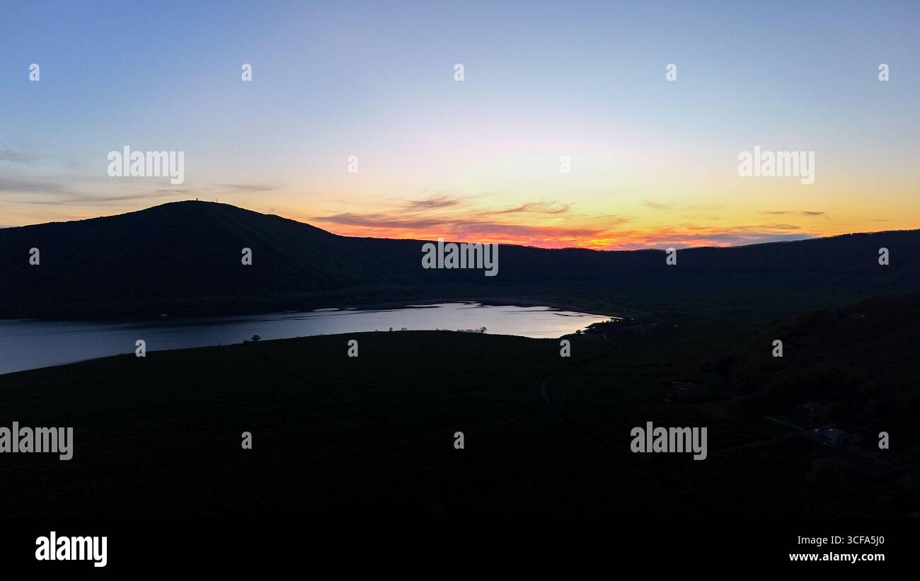I colori si fondono magnificamente mentre il sole tramonta dietro le colline, gettando riflessi sulle calme acque del lago. Un momento di pace nella natura, invitante e tranquillo Foto Stock