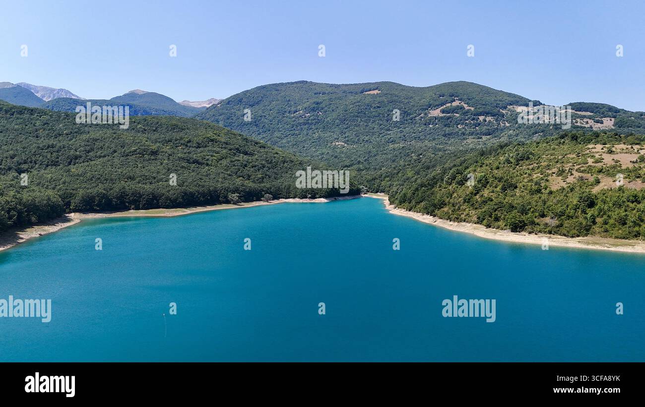 Sotto un radioso cielo di mezzogiorno, un tranquillo lago turchese riflette le maestose montagne circostanti. Alberi verdeggianti fiancheggiano la riva, creando un ambiente tranquillo Foto Stock