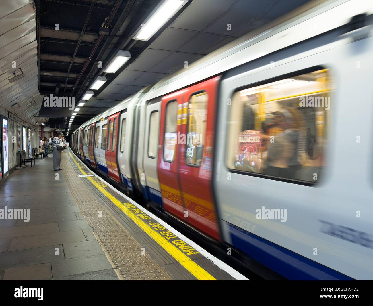 Un passeggero attende sulla piattaforma della metropolitana di Londra, mentre un treno con caratteristici telai rossi delle porte esce dalla stazione. Foto Stock