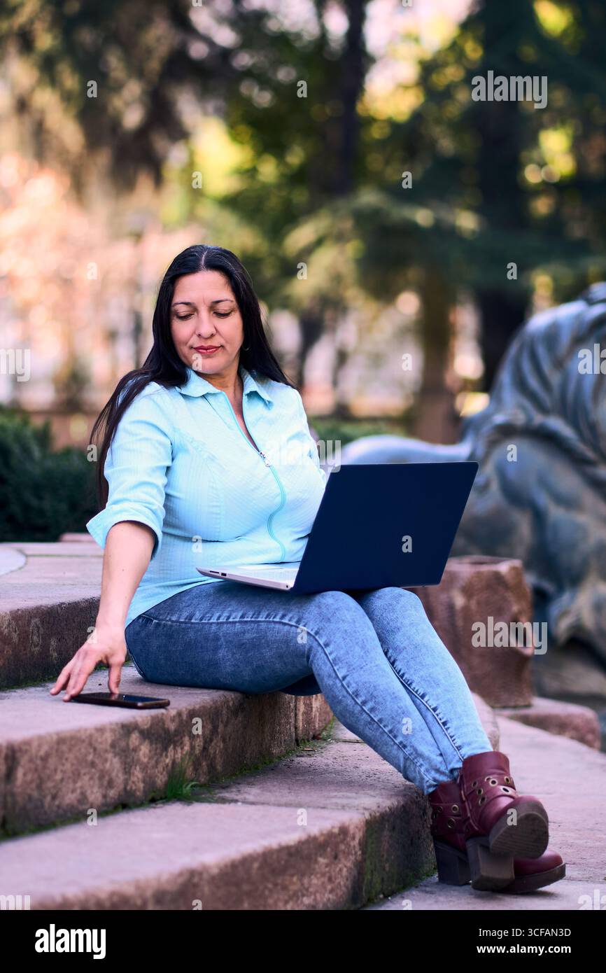 Bella donna latina matura che lavora sul suo portatile all'aperto a casa, sorride, mentre usa anche il suo cellulare. Donna d'affari, marketing, ufficio domestico. Foto Stock
