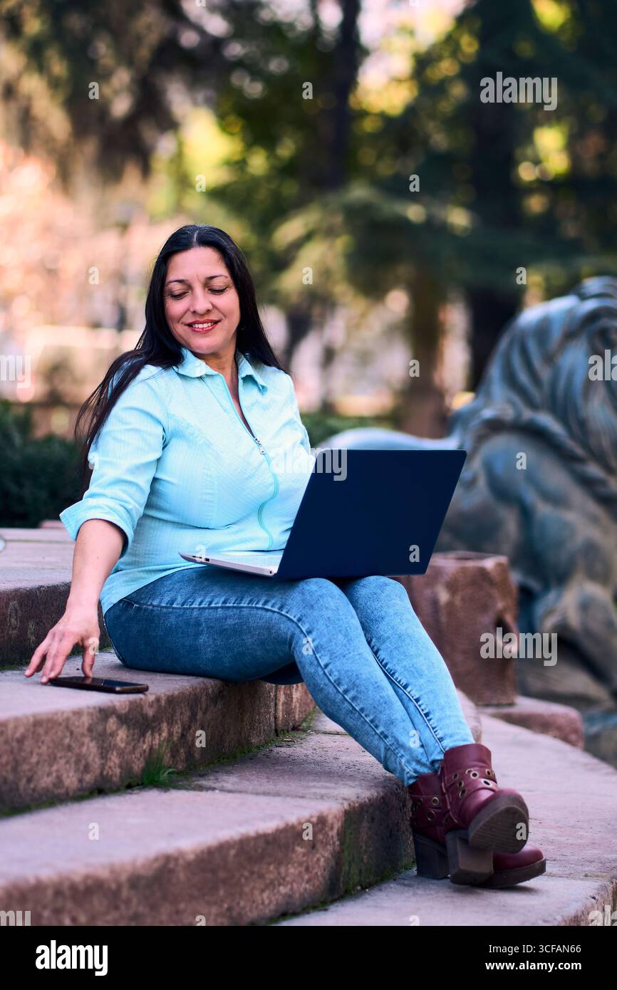 Bella donna latina matura che lavora sul suo portatile all'aperto a casa, sorride, mentre usa anche il suo cellulare. Donna d'affari, marketing, ufficio domestico. Foto Stock