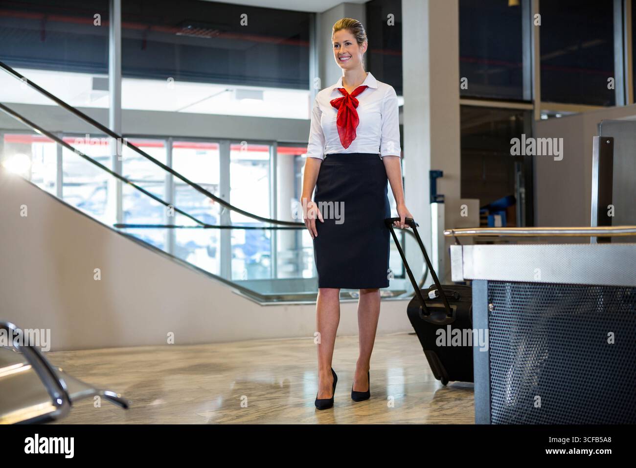 Donna assistente di volo in piedi e sorridente nel terminal che tiene una valigia con ruote vicino al banco del check-in Foto Stock