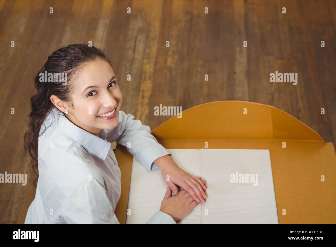 Donna che indossa una camicetta bianca seduta alla scrivania di legno sorridente alla macchina fotografica con un notebook aperto e spazio per le copie Foto Stock