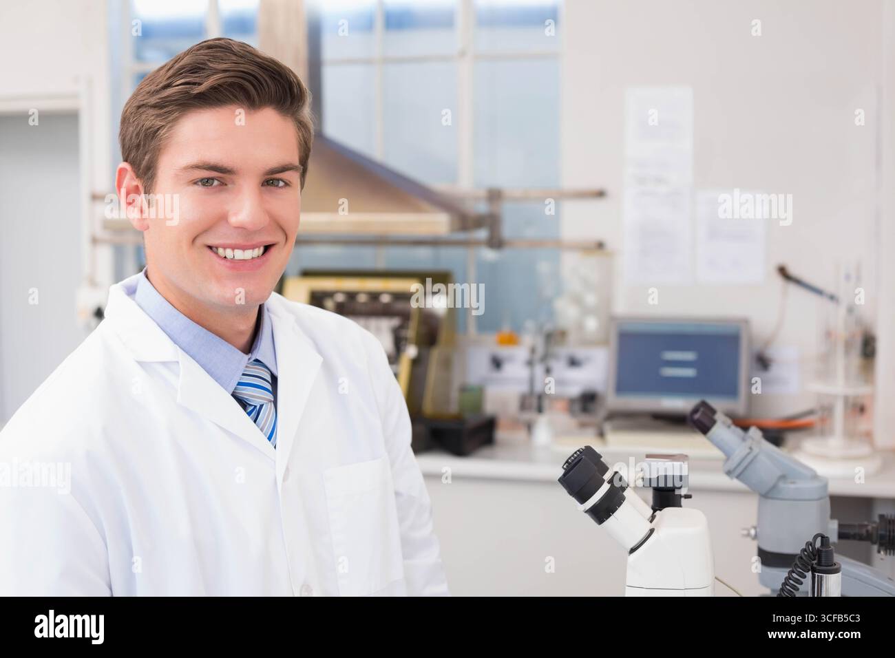 Ricercatore maschio in piedi in camice da laboratorio e cravatta sul moderno banco da laboratorio con microscopio, spazio di copia Foto Stock
