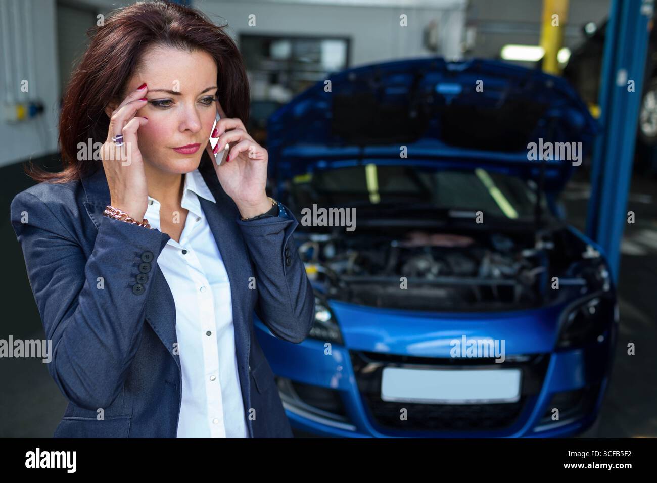 Donna che parla sullo smartphone accanto a un'auto blu con cofano aperto in officina, ascensore per veicoli, spazio fotocopie Foto Stock