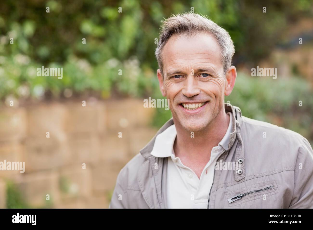 Uomo in piedi nel cortile posteriore che indossa una giacca grigio chiaro su una polo bianca con parete in pietra, spazio fotocopie Foto Stock