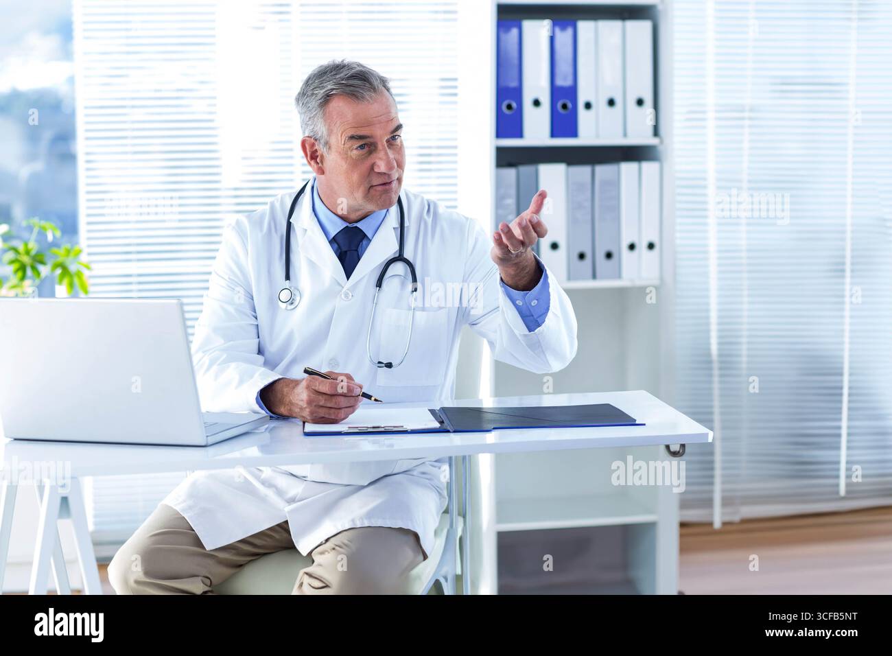 Medico di sesso maschile anziano che utilizza la penna sopra gli appunti alla scrivania, consultando il computer portatile e lo stetoscopio Foto Stock