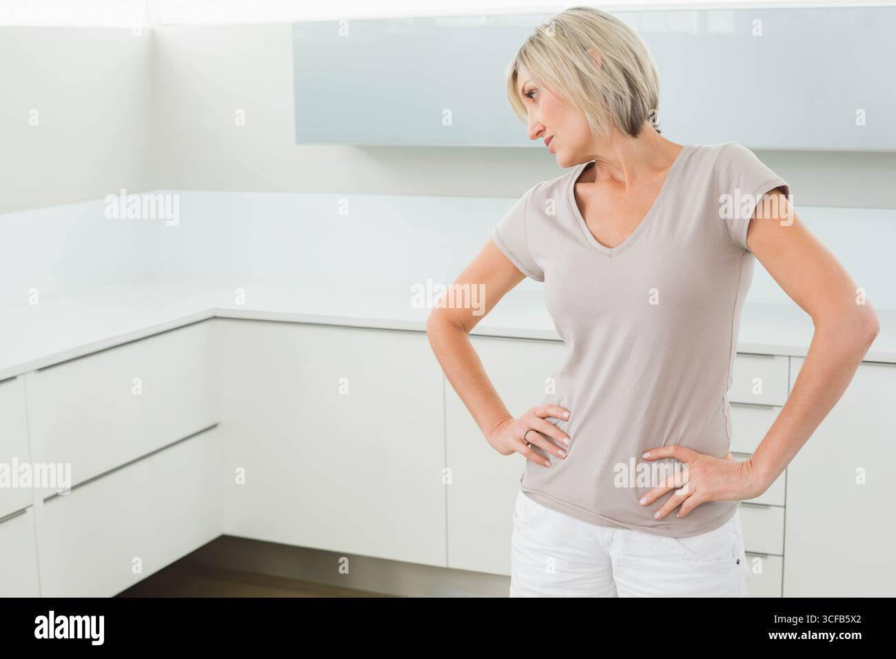 Donna di mezza età in piedi in una cucina moderna con armadi bianchi e piano di lavoro, spazio fotocopie Foto Stock