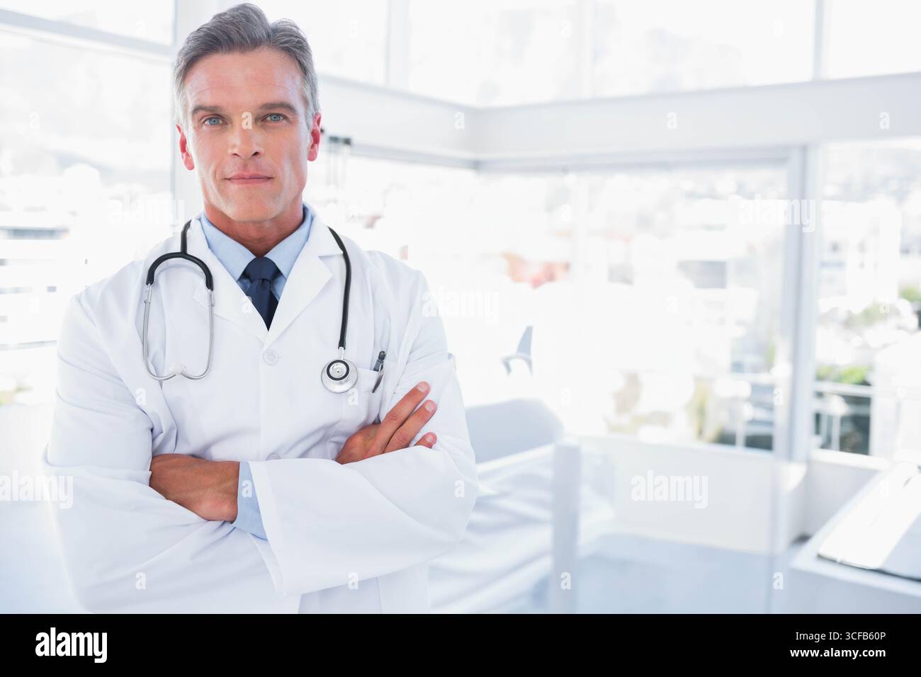 Medico maschio maturo in piedi con braccia piegate in clinica medica con stetoscopio, camice da laboratorio Foto Stock