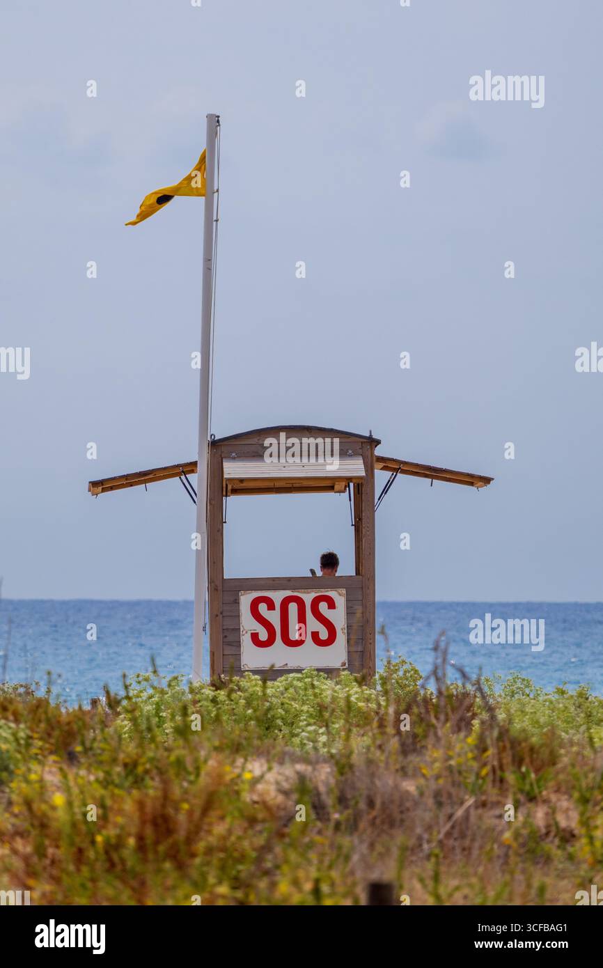 Bagnino che garantisce la sicurezza, osservando dalla torre del bagnino in legno con cartello sos e bandiera gialla che indicano condizioni del mare moderate Foto Stock