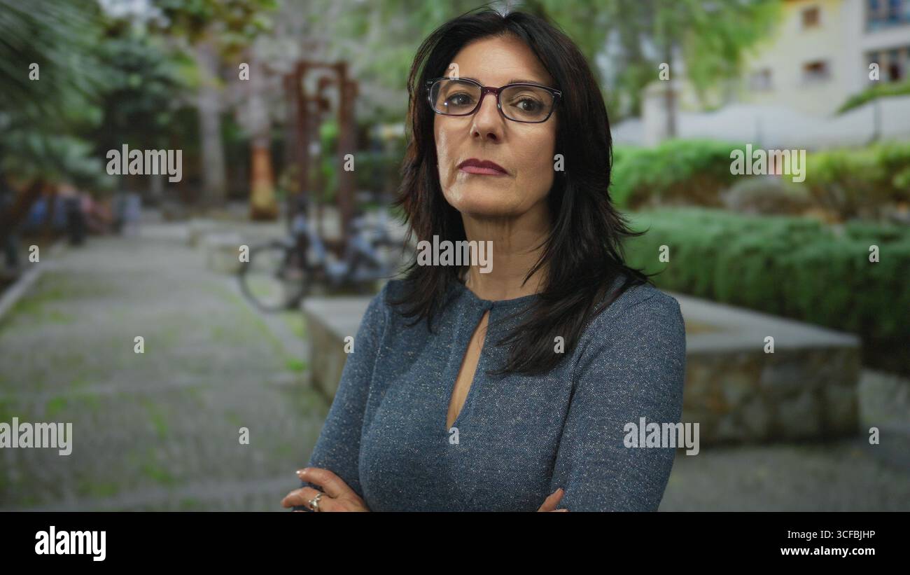 La donna ispanica che indossa gli occhiali sta con le braccia incrociate su un sentiero verde vicino a una panchina e a un portabiciclette; rifiuto. Foto Stock