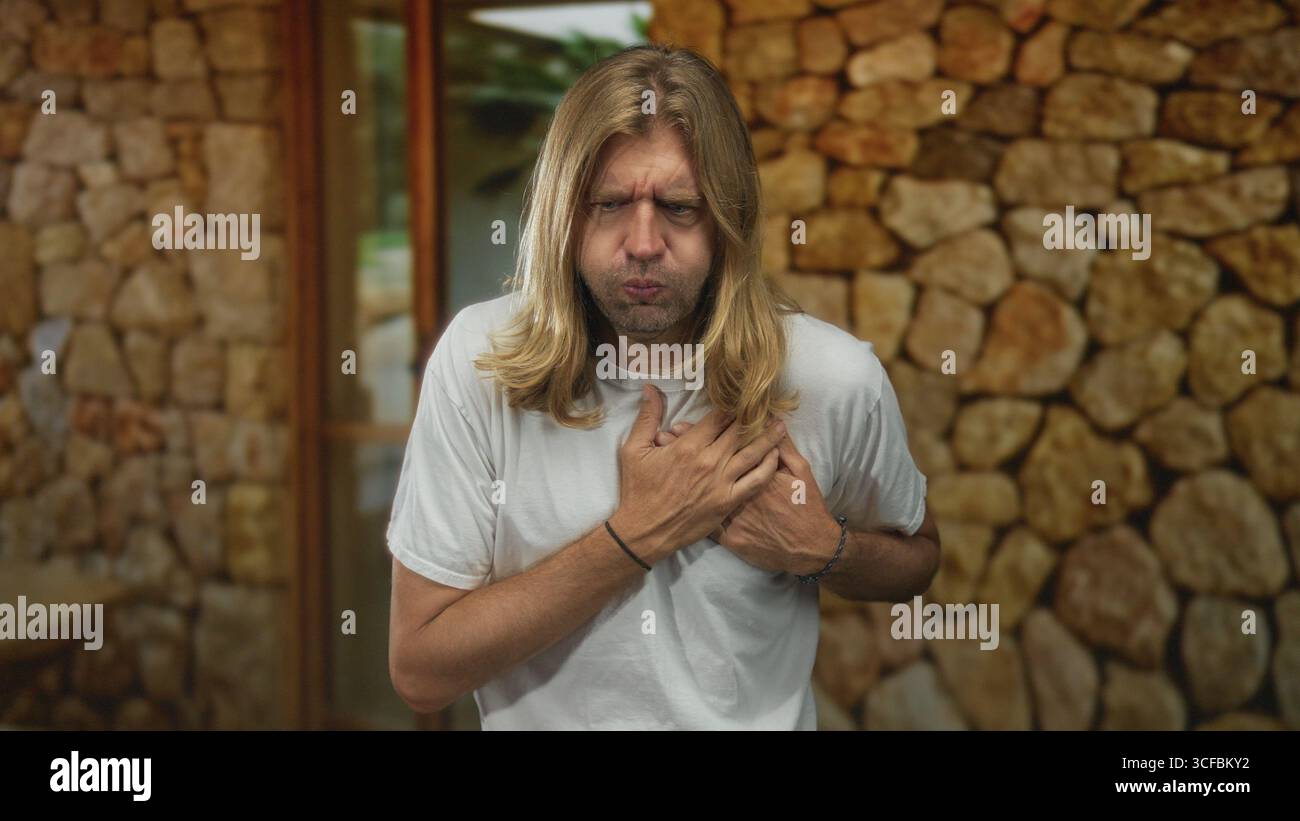 L'uomo biondo con i capelli lunghi stringe il petto per attacco di cuore mentre è in piedi alla porta di ingresso dell'edificio incorniciato da un muro di pietra; attacco di cuore vulnerabilità di soccorso Foto Stock