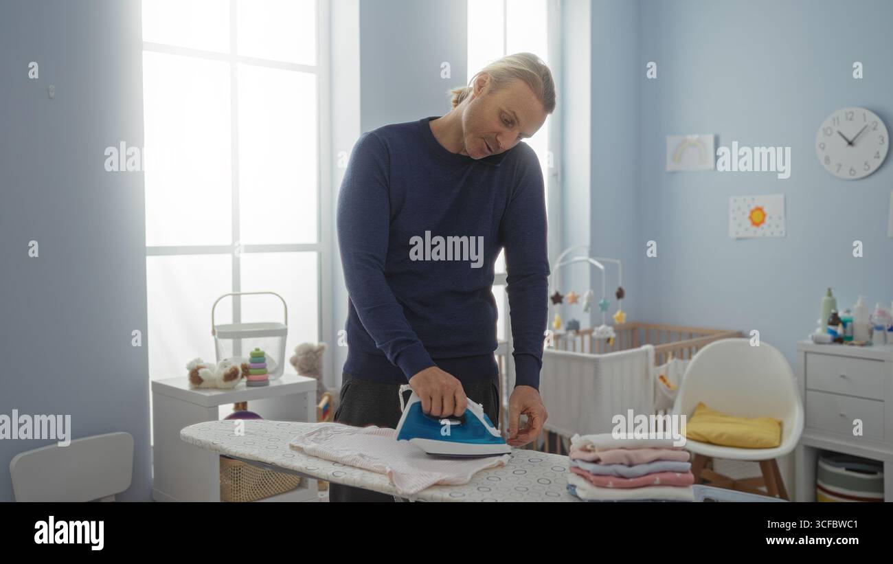 L'uomo maturo che stirava nella lavanderia interna di casa si concentra sulle faccende domestiche circondate da giocattoli per bambini che mettono in risalto la vita domestica e la responsabilità Foto Stock