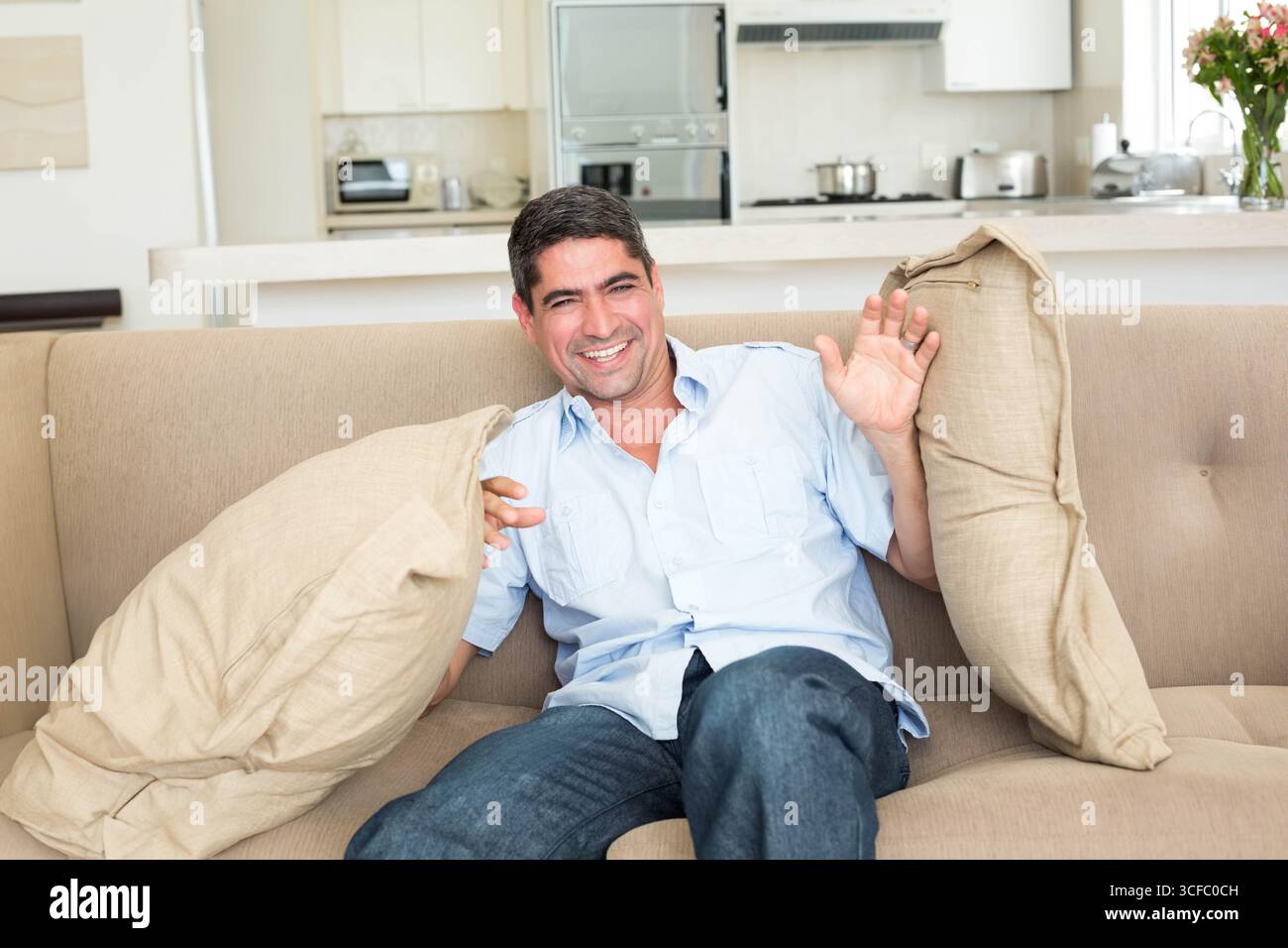 Uomo che ride tenendo i cuscini sul divano in soggiorno con elettrodomestici e fiori da cucina Foto Stock