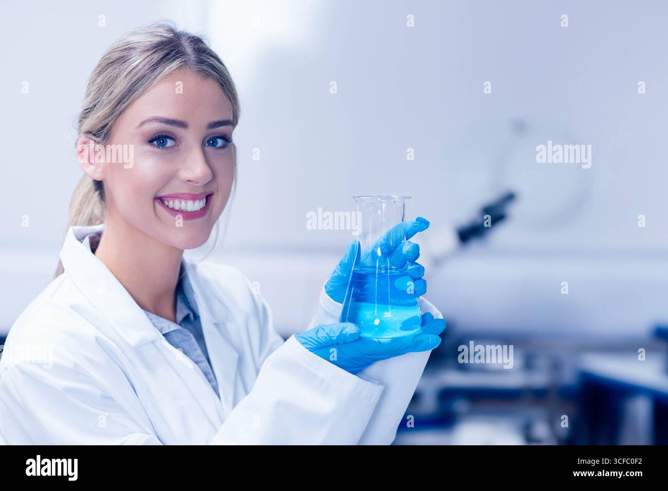 Scienziata donna che tiene in mano il becher con liquido blu indossando guanti in nitrile sul banco da laboratorio, spazio di copia Foto Stock