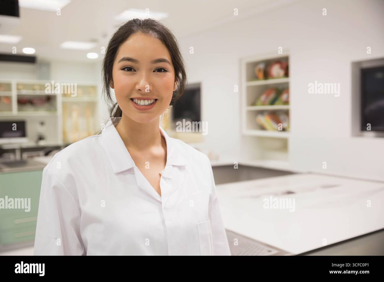 Scienziata asiatica sorridente in classe di laboratorio accanto a banchi di laboratorio e modelli di scheletro Foto Stock