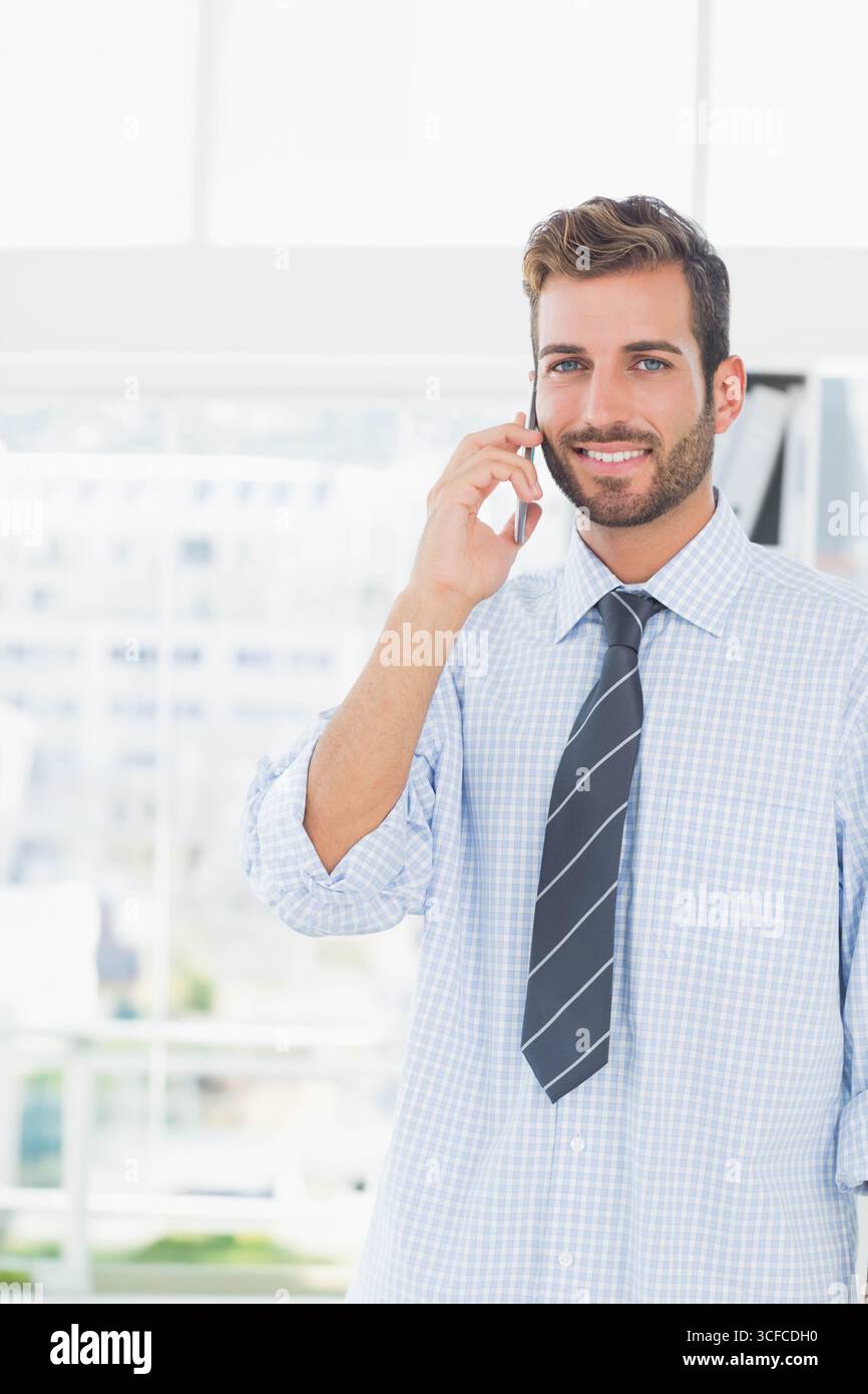 Uomo ispanico che indossa una cravatta accanto alla finestra dell'ufficio che tiene lo smartphone vicino ai raccoglitori e agli spazi per le copie Foto Stock