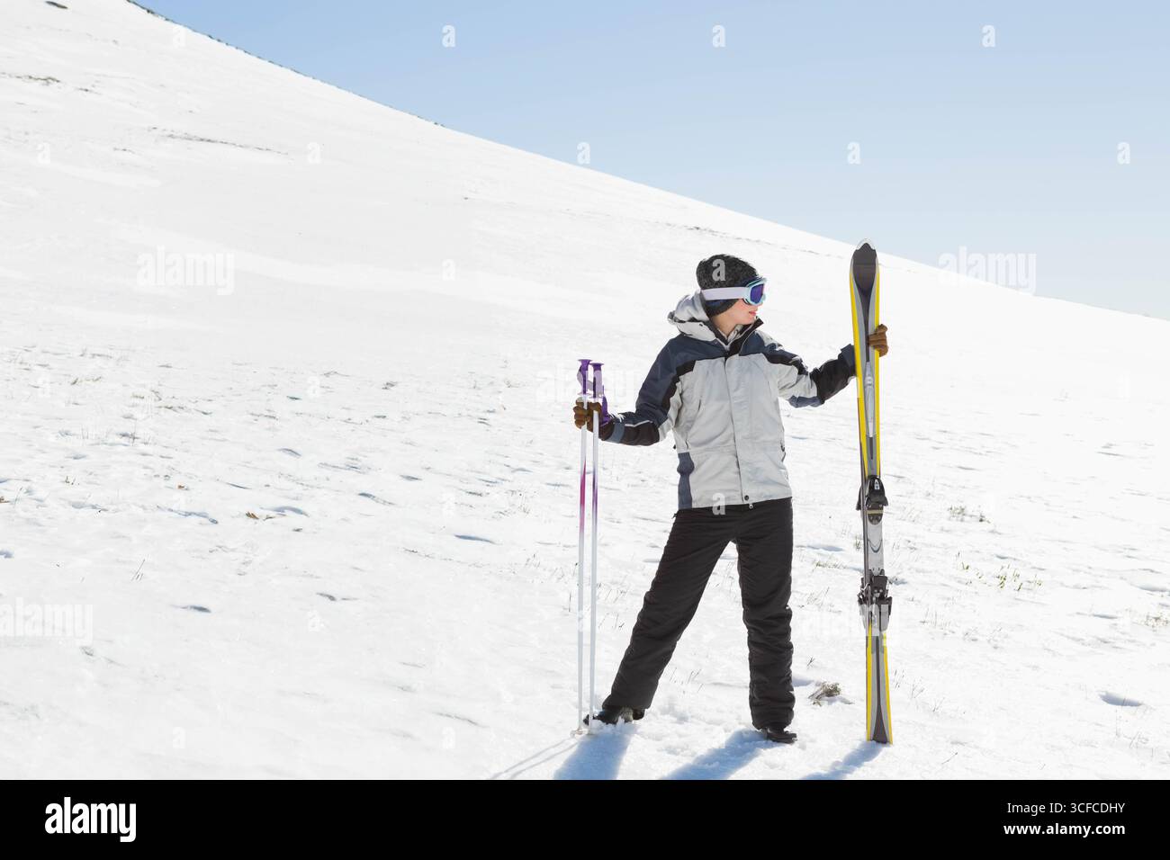 Sciatore in piedi su una collina innevata sotto il cielo azzurro, con sci gialli e pali viola Foto Stock