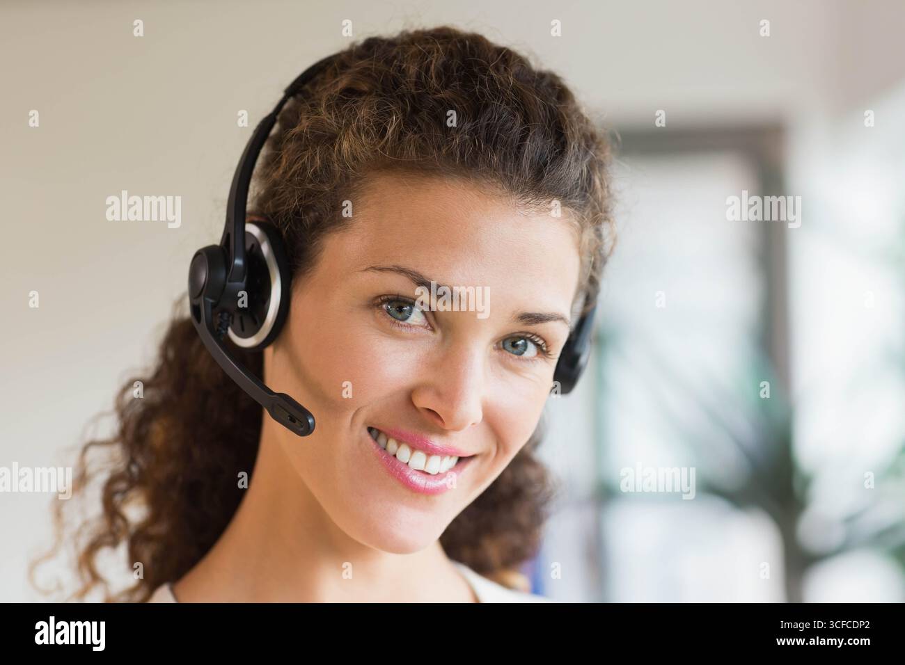 Donna che indossa l'auricolare e sorride alla finestra dell'ufficio con piante sfocate e pareti neutre Foto Stock