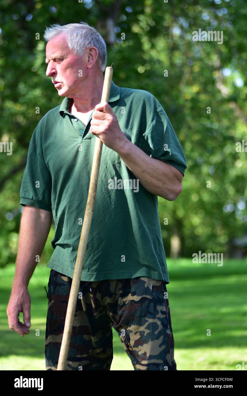 Ritratto ravvicinato di un uomo anziano con una camicia verde e pantaloni mimetici che tengono un bastone all'aperto, con un aspetto premuroso in un luminoso cortile di campagna wi Foto Stock