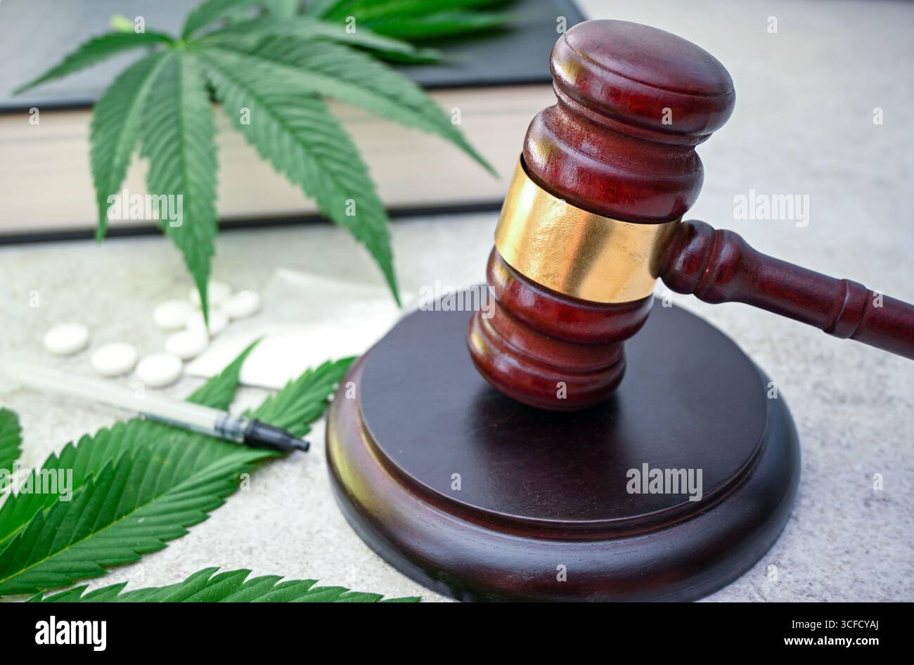 Il martelletto del giudice è seduto su un blocco sonoro con foglie di cannabis, pillole e siringa, simboleggiando la legalità della cannabis e i crimini legati alla droga. Foto Stock