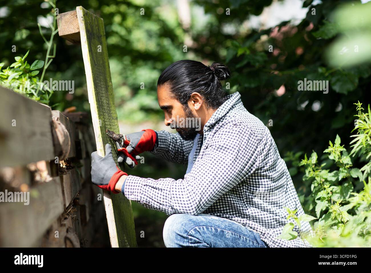 Un uomo si inginocchia all'aperto, misurando una tavola di legno con un metro a nastro in un lussureggiante giardino. Germania Foto Stock
