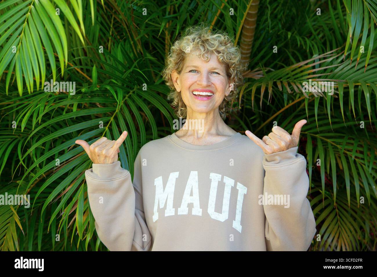 Donna sorridente in felpa beige con testo Maui, in piedi davanti a foglie verdi lussureggianti. Parco Kepaniwai, Wailuku, Hawaii Foto Stock