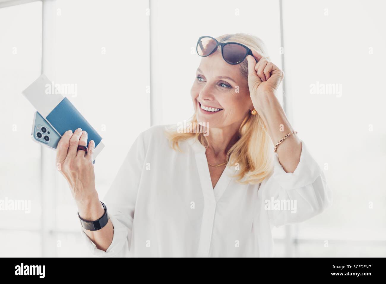 Donna matura in aeroporto che tiene in mano gli indispensabili per il viaggio, sorridendo e ammirando l'attesa del viaggio in estate Foto Stock