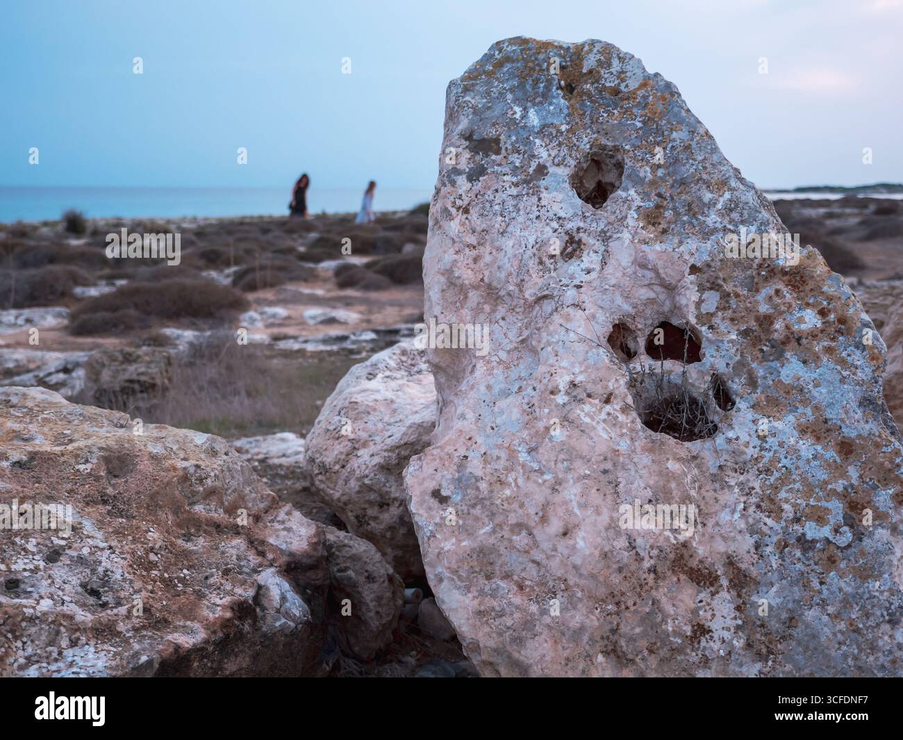 Primo piano di un'enorme roccia porosa con grandi buchi come impronte di zampe verticalmente sulla riva del Mar Mediterraneo, due donne turisti camminano lungo il mare Foto Stock