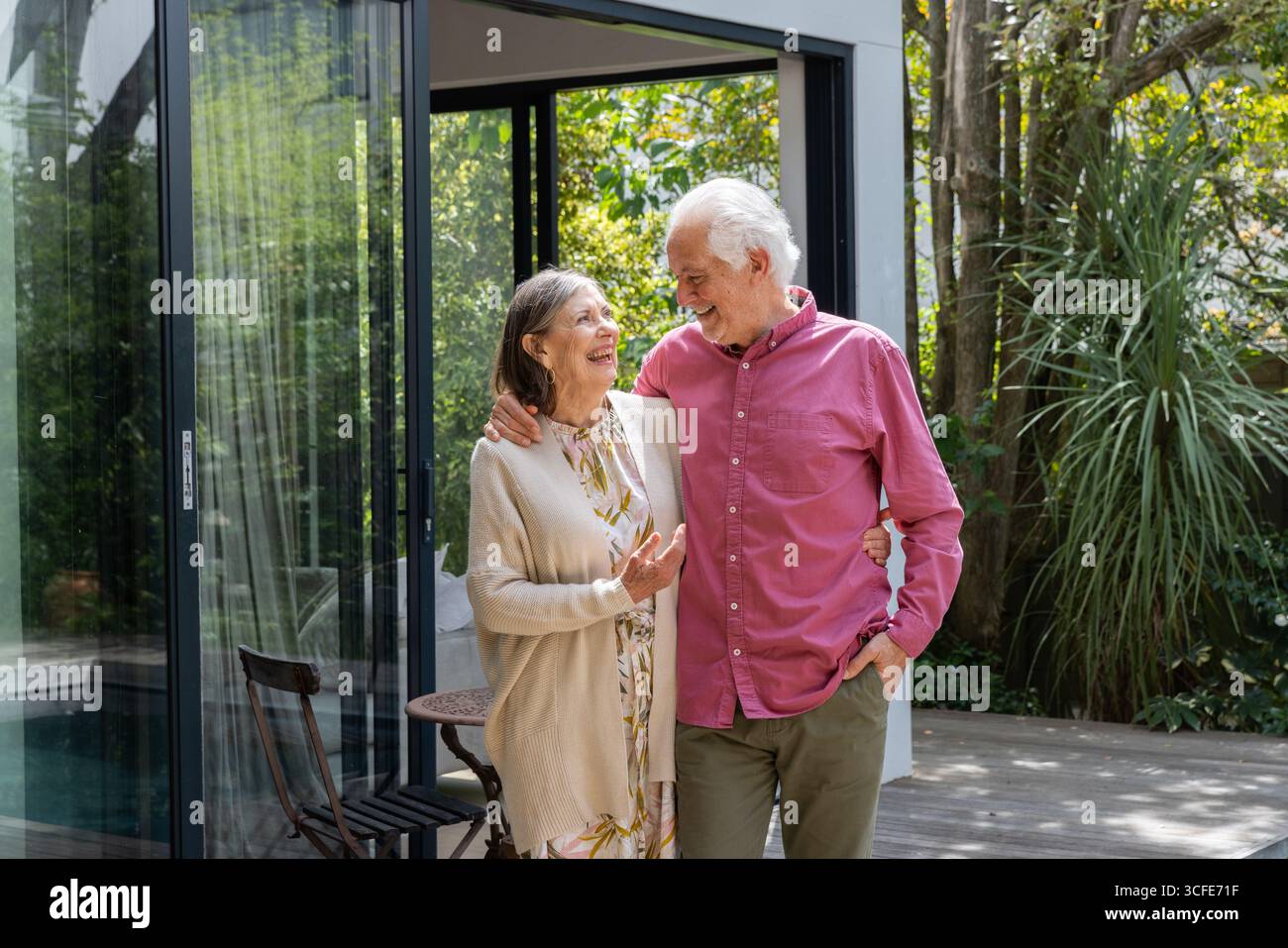 Una coppia anziana che cammina con il braccio sul patio in legno accanto a un piccolo tavolo rotondo fuori casa Foto Stock