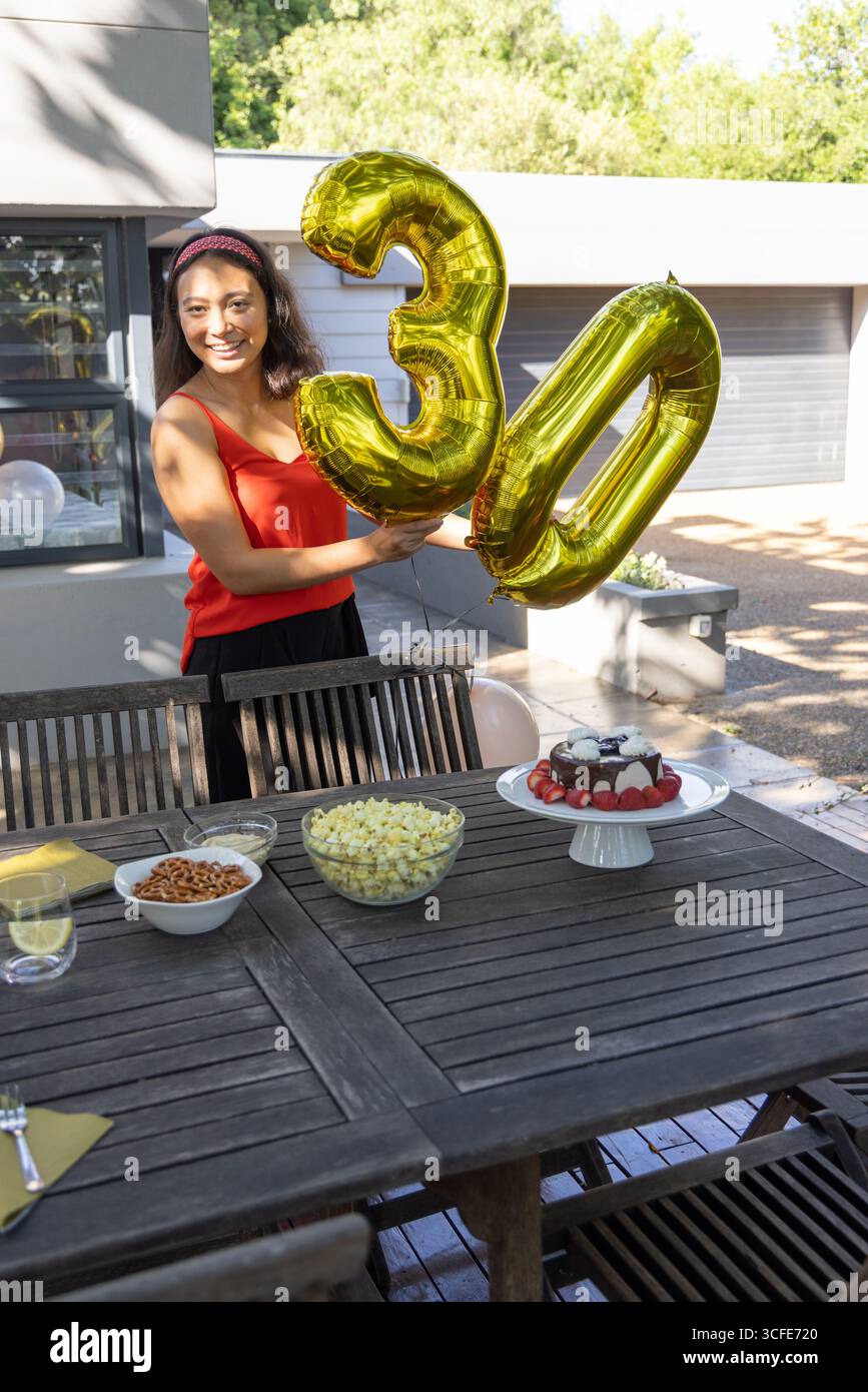 Donna asiatica in piedi dietro un tavolo di legno sul patio che regge palloncini d'oro 30, torta, spazio copia Foto Stock