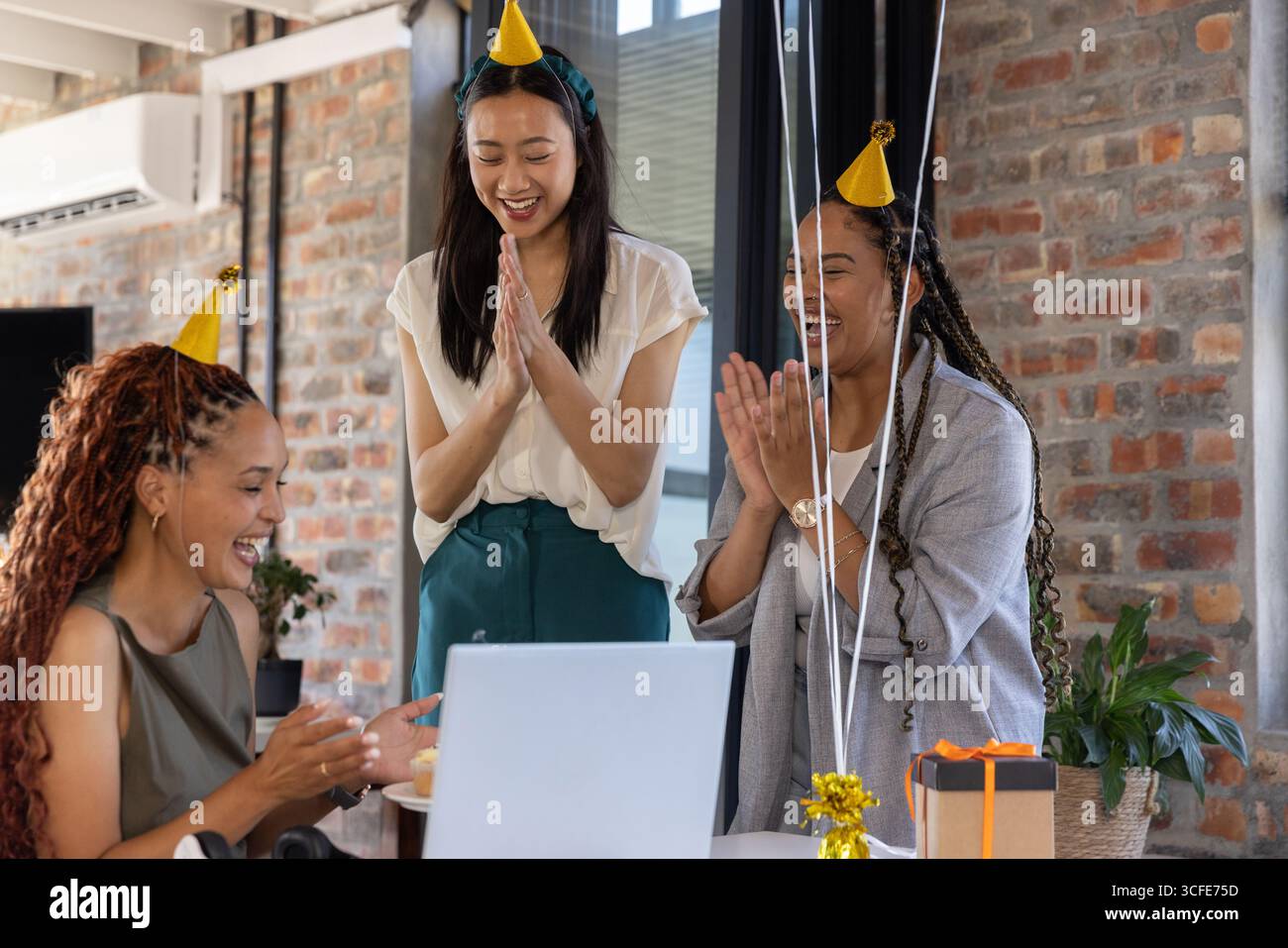 Diverse colleghe di cappelli da festa che applaudono e sorridono intorno al laptop con una confezione regalo in ufficio Foto Stock