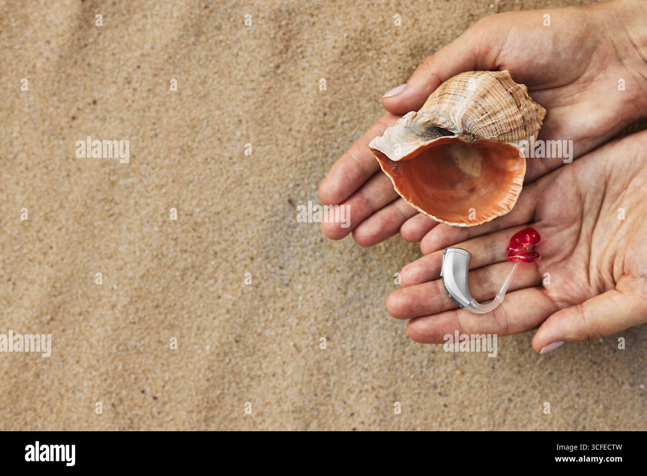 Le mani della persona tengono in mano un moderno apparecchio acustico e conchiglia sulla sabbia calda della spiaggia, simboleggiando la salute e il riposo Foto Stock