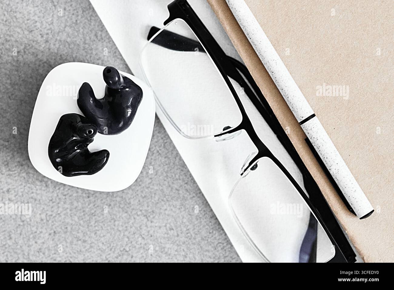 Coppia di tappi auricolari sagomati poggiano accanto agli occhiali e una penna su una scrivania minimalista, promuovendo soluzioni salutari e sonore, stile aziendale Foto Stock