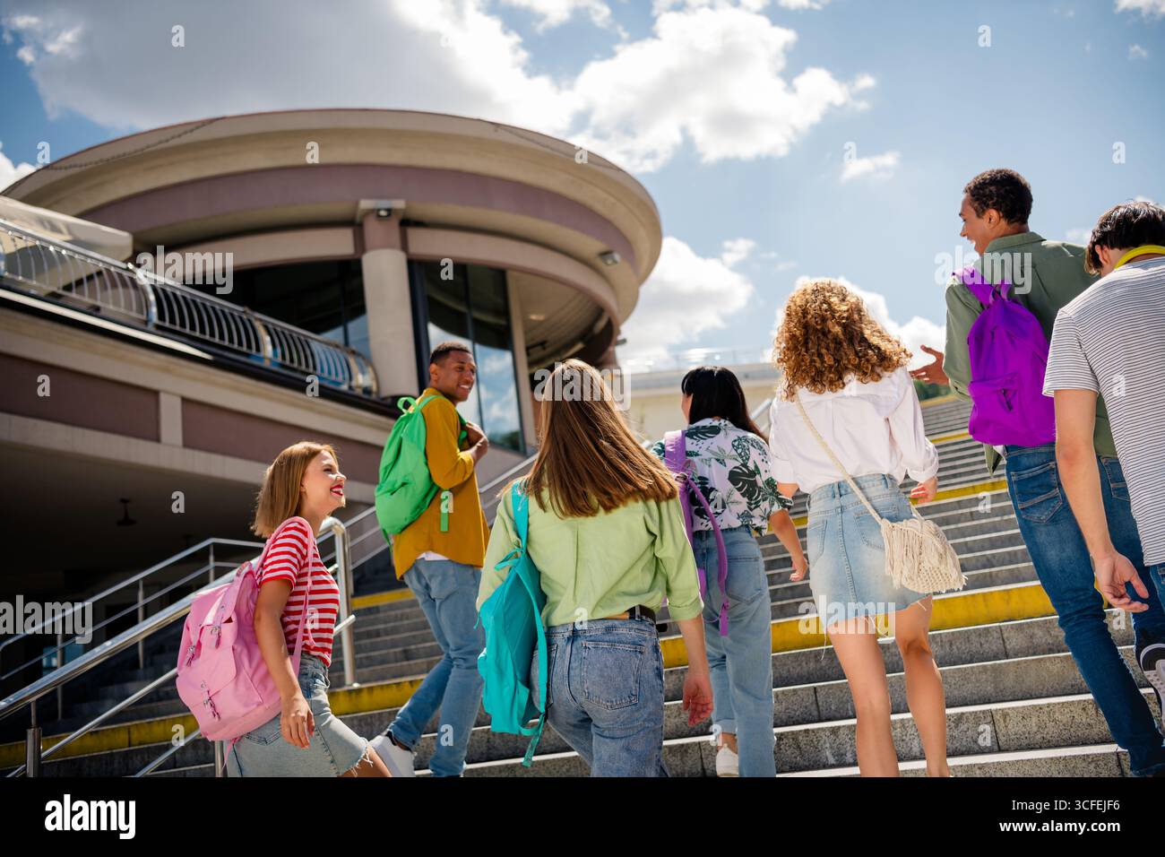 Gli studenti si godono una giornata di sole insieme salendo le scale per raggiungere un edificio moderno con zaini Foto Stock