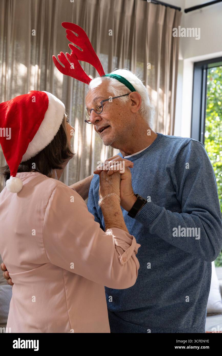 Coppia senior che tiene per mano e balla nel soggiorno con cappello di Babbo Natale e corna di renne Foto Stock