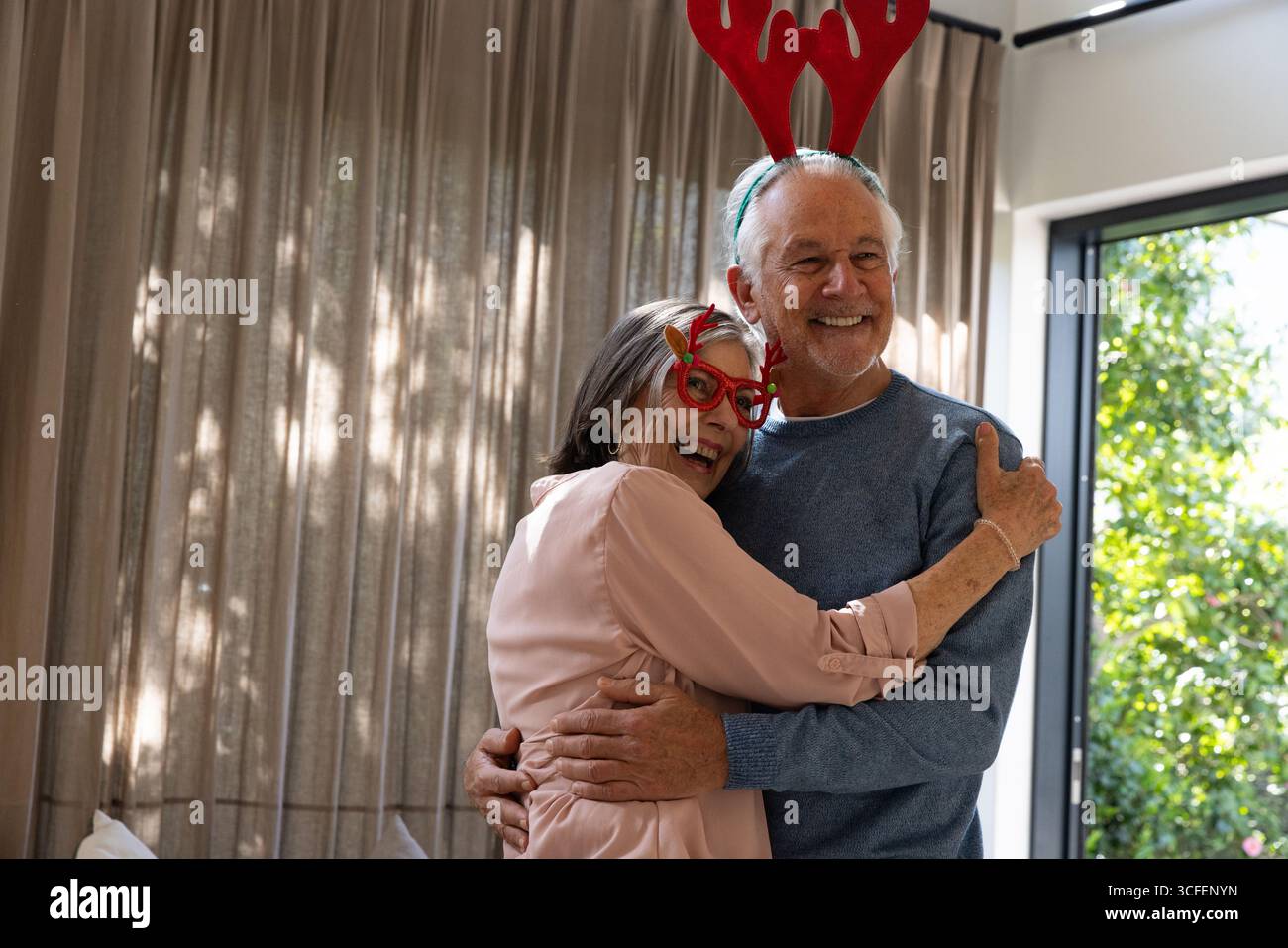 La coppia senior si abbraccia nel soggiorno indossando corna di renna e bicchieri nuovi vicino alle tende Foto Stock