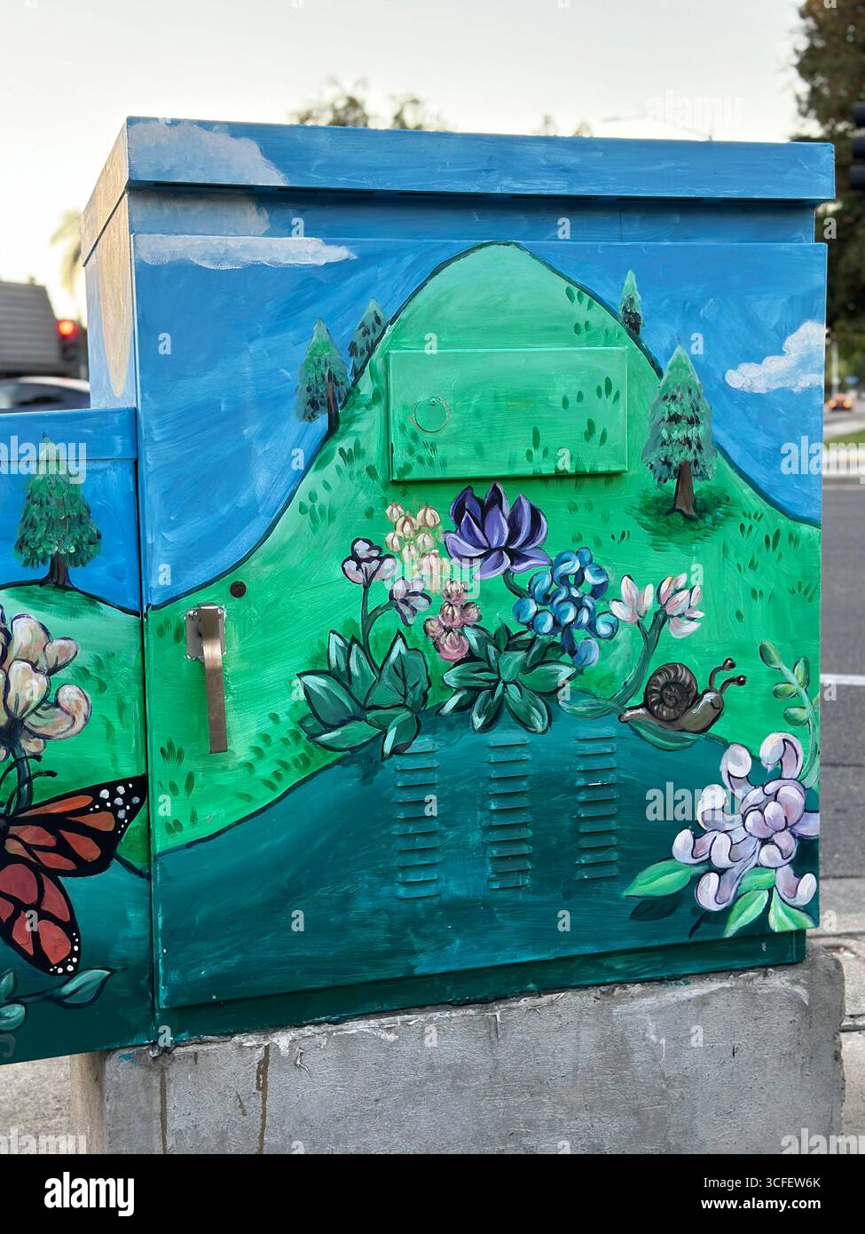 Una cassetta di servizio dipinta con colori vivaci che presenta una farfalla monarchica, fiori, colline verdi e cieli blu, catturati in un angolo di strada. - Immagine stock catturata con smartphone