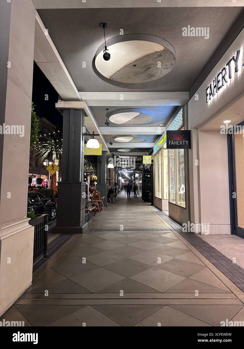Quartiere urbano dello shopping di notte con vetrine illuminate, palme e pedoni che camminano lungo la strada dei negozi all'aperto. - Immagine stock catturata con smartphone