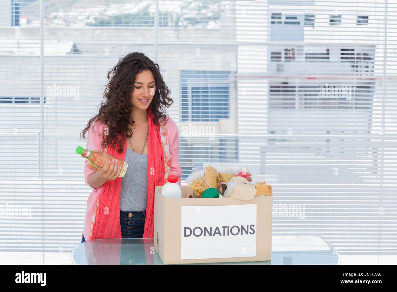 Volontaria donna che seleziona gli articoli alimentari in una scatola PER DONAZIONI da un tavolo di vetro nella stanza della comunità, copia spazio Foto Stock