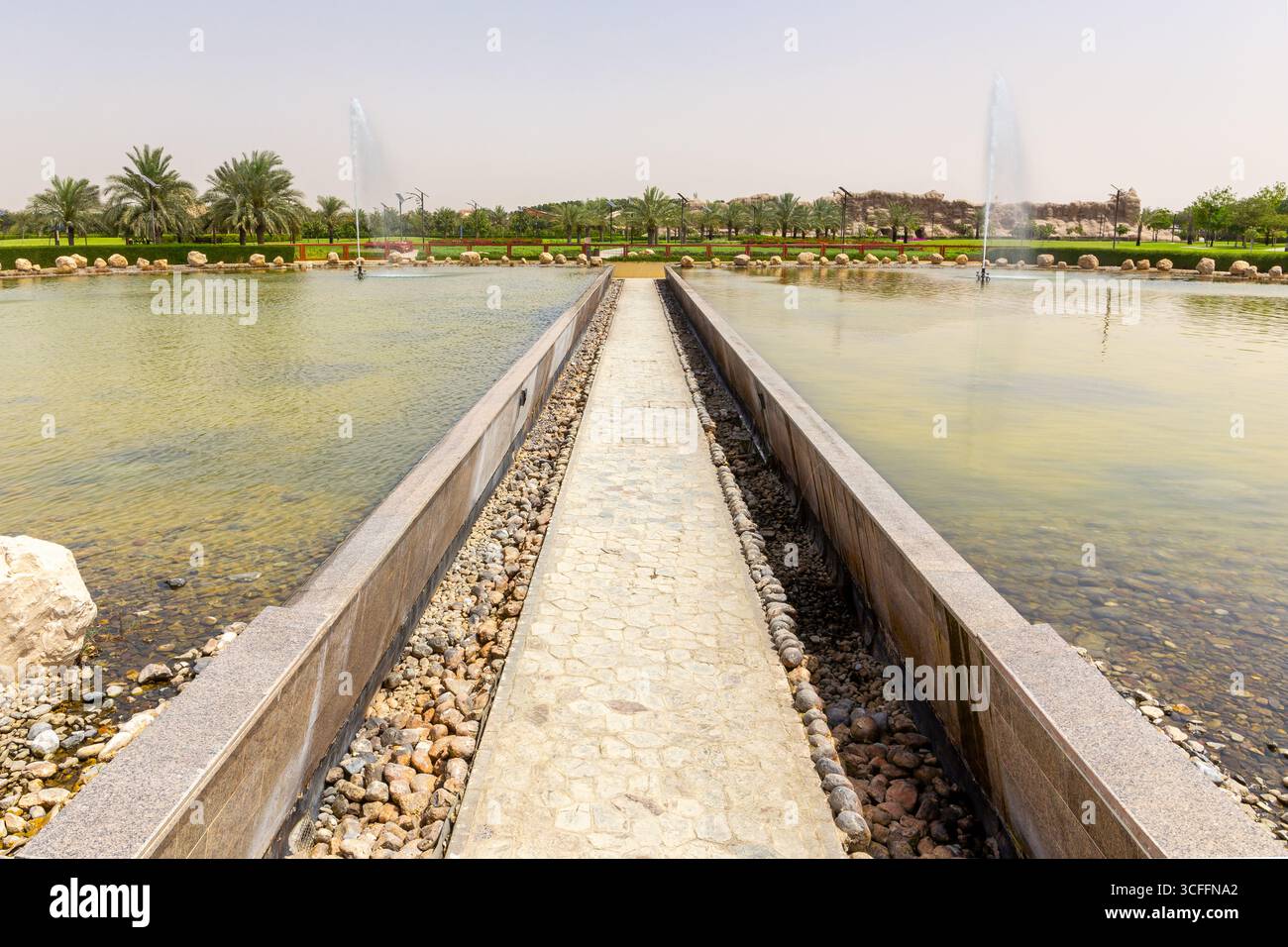 Sentiero a piedi del ponte di pietra attraverso il laghetto con fontane nel Parco del Corano, Dubai, Emirati Arabi Uniti, lussureggianti giardini verdi con palme da dattero. Foto Stock