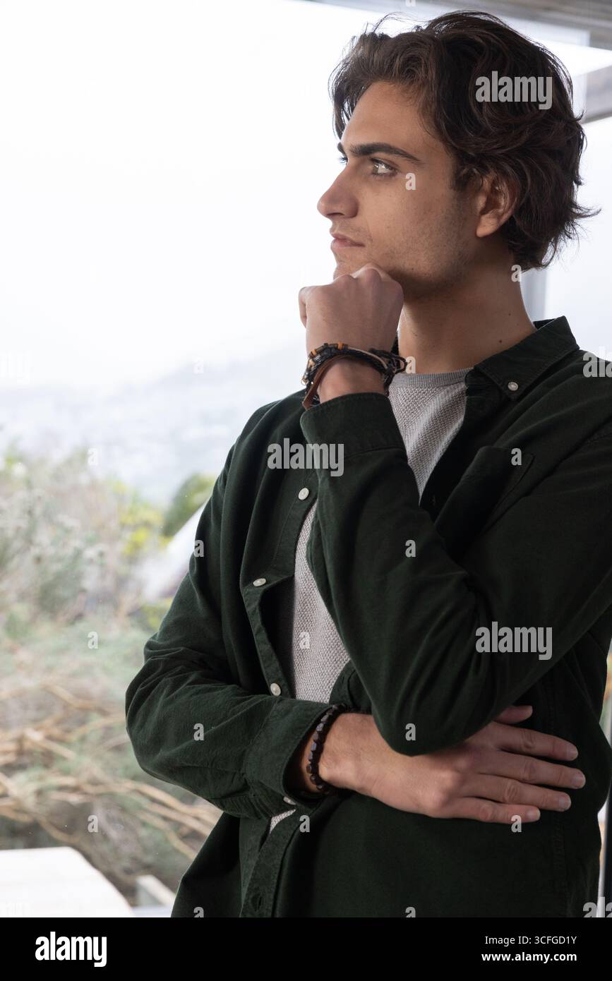 Uomo in piedi all'interno vicino a una grande finestra con indosso camicia e braccialetti, che guarda all'esterno, copia spazio Foto Stock