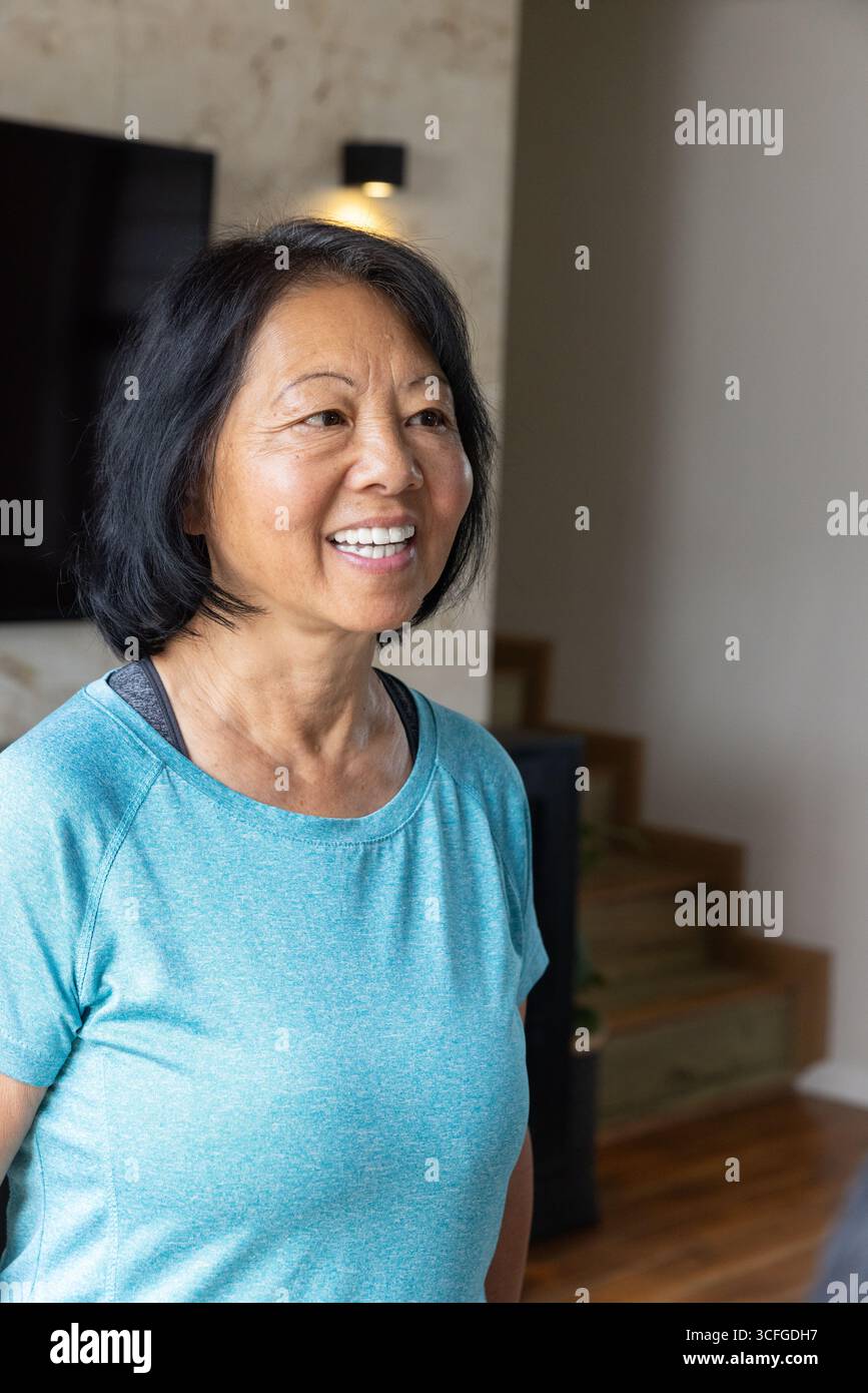 Donna asiatica anziana in piedi in un moderno salotto con top atletico e sorridente vicino alla televisione Foto Stock