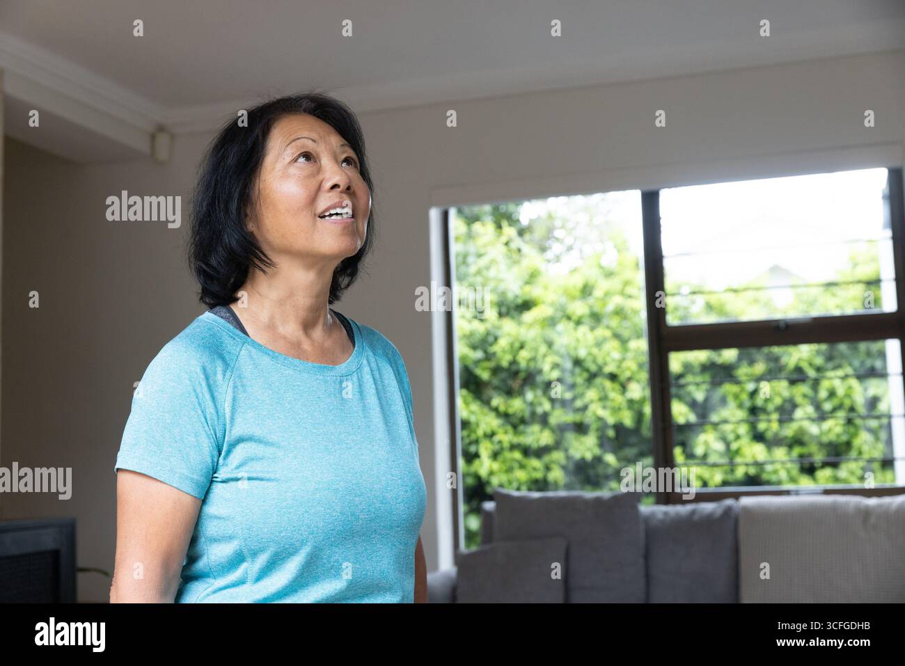 Donna asiatica anziana che indossa la parte superiore turchese che allunga il collo nel soggiorno vicino alla finestra, copia spazio Foto Stock